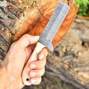 A hand holds the Texan Knives Gareth Bull Style Knife—8" Damascus steel blade, camel bone handle, and Western leather sheath—in front of a cut tree stump outdoors.