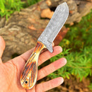 A hand holds the Whisky Bent Knives – Custom Handmade Damascus Steel Blade by Texan Knives, featuring a patterned blade and a textured brown and yellow camel bone handle, with blurred greenery and a log in the background.