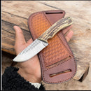 A person holds the Texan Knives Wrangler’s Edge Cowboy Knife with a patterned blade and stag horn handle, resting on its stitched brown leather case atop a wooden surface.