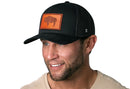 A smiling man with a short beard wears the Haka Hat Wyoming Flag Trucker Hat Leather in black, featuring a brown buffalo silhouette. He has on a beige shirt and stands against a white background.