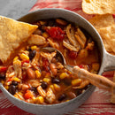 A bowl of chicken tortilla soup with beans, corn, tomatoes, and shredded chicken, seasoned with SALTOPIA Infused Sea Salts' EUREKA SWEET PAPRIKA (paprika & onion salt), garnished with a tortilla chip and served with extra chips. A spoon sits in the bowl.