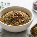 A white ceramic bowl holds SALTOPIA Infused Sea Salts' Saltopia Za'atar Spice Blend, featuring a mix of dried herbs and seeds, with a wooden spoon of the seasoning nearby and a glass jar in the background.