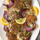 A platter of baked chicken drumsticks and thighs, seasoned with SALTOPIA Infused Sea Salts' Saltopia Za'atar Spice Blend, and garnished with fresh herbs, lemon wedges, and roasted red onion slices.