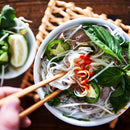A hand uses chopsticks to pick up noodles from a bowl of Vietnamese pho, garnished with basil, jalapeños, and sriracha. A sprinkle of The Spice Lab Birdseye Chili Pepper Powder by The Spice Lab adds a fiery kick to the aromatic broth. Nearby, a plate with lemon, herbs, and bean sprouts sits on a wooden table.