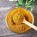 A glass bowl filled with vibrant yellow-orange The Spice Lab Indian Curry Seasoning Spice Maharaja Style, celebrated for its full-bodied curry flavor, with a wooden spoon resting inside, set atop a wooden surface and green leaves in the background.
