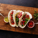 Three steak tacos with cilantro, grilled vegetables seasoned with The Spice Lab Organic Ground Cumin, and pickled onions on a wooden board, served with green salsa, red salsa, and sliced jalapeños on the side, shot from above.
