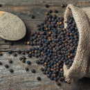 A burlap sack spills whole organic peppercorns onto a rustic wooden surface, while a wooden spoon holds The Spice Lab Organic Ground Black Pepper nearby.