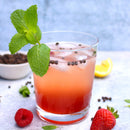 A pink iced drink garnished with mint leaves sits in a glass, topped with The Spice Lab Special Extra Bold Indian Black Peppercorns, and is surrounded by strawberries, raspberries, a lemon wedge, and mint on a light surface.