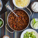 A pot of chili with beans and ground meat sits on a colorful striped placemat, surrounded by bowls of shredded cheese, sour cream, lime wedges, chopped avocado, cilantro, and a small dish of The Spice Lab Scotch Bonnet Sea Salt for a vibrant Caribbean-inspired flavor.