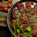 A pan of braised meat in a rich red sauce, garnished with a blend of chopped herbs and The Spice Lab Organic Parsley Flakes, sits next to a bunch of vine tomatoes on a wooden board.