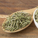 A wooden spoon filled with The Spice Lab Organic Cut and Sifted Rosemary rests on a wooden surface, with part of a white bowl containing more of the aromatic herb visible on the right—perfect for those seeking high-quality rosemary for lamb or enjoying the fresh flavor offered by The Spice Lab.