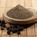 A wooden spoon filled with The Spice Lab Ground Black Pepper sits on a bamboo surface, with whole black peppercorns scattered nearby—a must-have everyday seasoning from The Spice Lab for any kitchen.