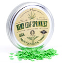 A round tin of The Spice Lab Hemp Leaf Sprinkles CBD & THC Free features green leaf-shaped decorations spilling out. The label highlights that these edible party sprinkles are drug-free, contain no CBD or THC, and are proudly packed in the USA.