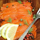 Thin slices of smoked salmon on a wooden platter, garnished with fresh dill, lemon slices, capers, and The Spice Lab Kings Blend Peppercorn Grinder Refill from The Spice Lab, with a fork on the side.