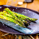 Grilled asparagus spears topped with coarse pepper and a sprinkle of The Spice Lab Fleur de Sel Sea Salt by The Spice Lab, served on a dark ceramic plate with a decorative blue brushstroke, atop a wooden table.