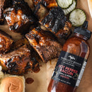 A bottle of Terrapin Ridge Farms Hot Pepper Bacon Grill & Wing Squeeze is next to saucy grilled ribs, pickle slices, and a piece of bread on a parchment-lined surface.