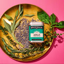 A gold plate on a pink background holds fresh herbs—sage, basil, rosemary, thyme, lavender—and a tin of Spicewalla Herbs de Provence by Spicewalla, with some loose dried lavender scattered on the plate.