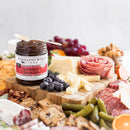 A charcuterie board with cheese cubes, berries, gherkins, meats, nuts, grapes, and a jar of Terrapin Ridge Farms Hot Pepper Bacon Jam. Background is softly blurred. Perfect for tasting from the 1 Gallon Jug by Terrapin Ridge Farms.