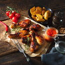 Grilled chicken drumsticks on parchment are arranged with rosemary, cherry tomatoes, potato wedges, a red chili, jarred sauce, a pepper grinder, and spices—including The Spice Lab Organic Minced Garlic—on a rustic wooden table.