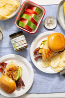 Two plates with chicken sandwiches on buns, each with bacon, avocado, and cheese, served with chips. A bowl of chips and watermelon slices pair perfectly with Terrapin Ridge Farms’ Amber Ale Pineapple Jalapeño Jam for a sweet heat kick.