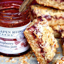 A close-up of oat bars filled with Terrapin Ridge Farms Raspberry Amaretto Jam. One bar leans against an open jar of the Terrapin Ridge Farms jam, a wooden spoon inside, with crumbs scattered around these gourmet treats.