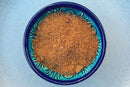 A blue ceramic bowl holds a serving of brown, granulated palm sugar topped with a sprinkle of World Spice Anardana from World Spice, sitting on a light gray surface and adding a subtle hint of tangy spice to the scene.