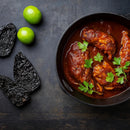 A black skillet filled with pieces of chicken in red sauce, garnished with fresh parsley, exudes authentic Mexican flavor. Surrounding the dish are two whole limes and two Rico Rico Ancho Dried Chiles – Sweet & Smoky Mexican Peppers (Mild Heat) from Rico Rico, all arranged on a dark surface.