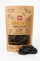 A brown resealable bag labeled Rico Rico Ancho Dried Chiles – Sweet & Smoky Mexican Peppers (Mild Heat) shows dried poblano peppers inside with some displayed in front. The Rico Rico packaging features illustrations of chiles, notes mild heat, and promises authentic Mexican flavor.