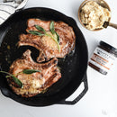 Two cooked pork chops in a cast iron skillet, garnished with fresh sage, are served alongside mashed potatoes and an open quart jar of Terrapin Ridge Farms Apple Maple Bacon Jam on a white surface, ready to add sweet and savory flavor.