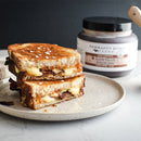 A grilled cheese sandwich with melted cheese and bacon, cut in half and stacked on a beige plate, sits in front of a jar of Terrapin Ridge Farms Apple Maple Bacon Jam Quart on a marble surface.