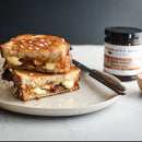 A grilled cheese sandwich with melted cheese and bacon sits on a plate with a knife beside it. In the background is a 5oz jar of Terrapin Ridge Farms Apple Maple Bacon Jam, perfect for charcuterie boards.