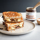 A grilled cheese sandwich, halved and stacked on a plate, oozes melted cheese and savory bacon jam. Coarse salt is sprinkled on top, with a jar of Terrapin Ridge Farms Apple Maple Bacon Jam in the background.
