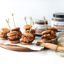 Mini chicken and waffle sliders topped with sauce are served on parchment, ideal for charcuterie boards. Jars of Terrapin Ridge Farms Apple Maple Bacon Jam and a basting brush are nearby for serving.