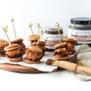 Mini fried chicken and waffle sliders are served on a wooden board, each with a skewer. Jars of spice maple syrup and Terrapin Ridge Farms Apple Maple Bacon Jam Quart bring extra flavor, ideal for an inviting brunch or appetizer spread.
