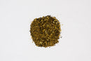 A small pile of World Spice Asian Tea Rub, a mixed spice blend from World Spice featuring green, yellow, and brown flecks, sits on a plain white background—hinting at its intriguing smoky flavor inspired by Lapsang Souchong tea.