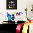 A collection of awards and trophies—including glass sculptures, metal statues, colorful ribbons, and plaques—stands beside The Spice Lab Ultimate Organic Spice Set - 24 Jars, a USDA Certified Organic product, showcasing The Spice Lab’s commitment to exceptional quality and outstanding achievements.