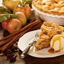 A slice of apple pie topped with a scoop of vanilla ice cream and drizzled with caramel sauce sits on a plate beside cinnamon sticks, fresh apples, spices, and a whole pie in the background—evoking the warm, comforting aroma of The Spice Lab Apple Pie Spice Blend by The Spice Lab, the perfect addition to classic baked treats.