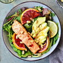 Enjoy a vibrant salad featuring mixed greens, a grilled salmon fillet, avocado slices, blood orange segments, and a sprinkling of The Spice Lab Organic Pine Nuts from The Spice Lab as a flavorful topping, served with a lime wedge in a gray bowl on a textured surface.