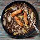 Three browned sausages sit atop cooked lentils, leeks, and The Spice Lab Organic Fennel Seeds in a black skillet with a metal serving spoon, set on a rustic wooden surface. Enhanced by the aromatic fennel seeds from The Spice Lab, the dish offers an inviting aroma and a mild licorice flavor—perfect for those who love cooking and baking with natural ingredients.