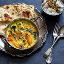 A bowl of yellow curry with prawns and vegetables, simmered with aromatic spice blends including The Spice Lab Organic Ground Mustard, is served on a silver plate alongside naan. Nearby sits a small bowl of rice garnished with fried onions and herbs, accompanied by a fork and spoon.
