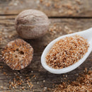 A whole nutmeg, a halved nutmeg, and a white spoon filled with freshly grated nutmeg from The Spice Lab Whole Nutmeg rest on a rustic wooden surface, with some grated nutmeg scattered around. Sourced from Indonesia, The Spice Lab brings a warm, aromatic touch to any dish.