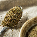 A close-up of a spoon filled with The Spice Lab Whole Celery Seeds, resting on a beige cloth beside a ceramic bowl containing more seeds—perfect for adding savory flavor or blending with pickling spices in your favorite recipes.