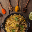 A pot of spiced rice with chicken, seasoned with The Spice Lab Ground Coriander 1 lb Bulk Foodservice Spice and garnished with cilantro, is surrounded by wooden spoons holding colorful spices and fresh cucumber slices on a rustic wooden table—a feast reminiscent of signature dishes found in BBQ restaurants.