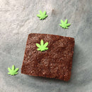 A chocolate brownie topped with a green hemp leaf-shaped decoration made from The Spice Lab Hemp Leaf Sprinkles CBD & THC Free by The Spice Lab, surrounded by three similar edible party decorations on a gray surface.