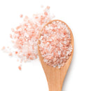 A wooden spoon filled with coarse Himalayan Pink Salt crystals, with some salt scattered on a white background—a perfect complement to The Spice Lab Himalayan Pink Salt & Premium Kings Pepper Grinder Set by The Spice Lab.