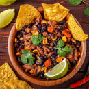 A wooden bowl of chili made with black beans, corn, carrots, and ground meat—seasoned with The Spice Lab Organic Chili Powder and topped with cilantro and a lime wedge—surrounded by tortilla chips on a wooden table.