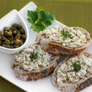 Three slices of seeded bread topped with a creamy spread and a sprinkle of The Spice Lab Fancy Chervil are arranged on a white plate next to a small dish of capers and a sprig of parsley—an elegant yet simple tribute to classic French cuisine, featuring the fine herbs from The Spice Lab.
