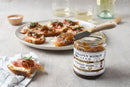 A quart jar of Terrapin Ridge Farms Balsamic Onion Jam with a knife on top sits beside slices of bread topped with the jam and prosciutto—ideal for charcuterie boards, paired with wine glasses and a napkin in the background.