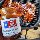 A hand holds a jar of DDR BBQ Supply Bacon Bourbon BBQ Sauce – 16 oz in front of a grill, where ribs glazed with the smoky sauce sizzle over glowing charcoal.