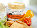 An open jar of Terrapin Ridge Farms Hatch Chile Bacon Ranch Dip sits surrounded by colorful bell peppers and crispy chips, offering a smoky southwest flavor, while another dip jar from Terrapin Ridge Farms is partially visible beside it.